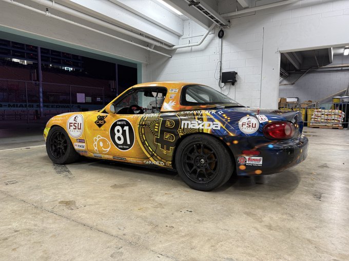 A yellow and blue Mazda Miata race car with the number 81, featuring sponsors including FSU, mxpa, Da, Akrapovic, and CRS, parked in what appears to be a Miami Speedway garage.