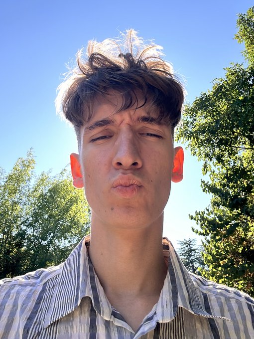 A close-up selfie of a young man with wavy brown hair squinting in bright sunlight, wearing a striped shirt, with green trees and blue sky visible in the background.