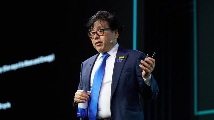 A man in a dark suit with a blue tie and a yellow pin is speaking at an event, holding a clicker, with text partially visible in the background.