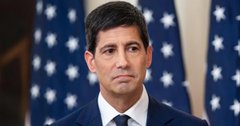 A portrait of a man with short dark hair wearing a dark suit and blue tie, standing in front of American flags.