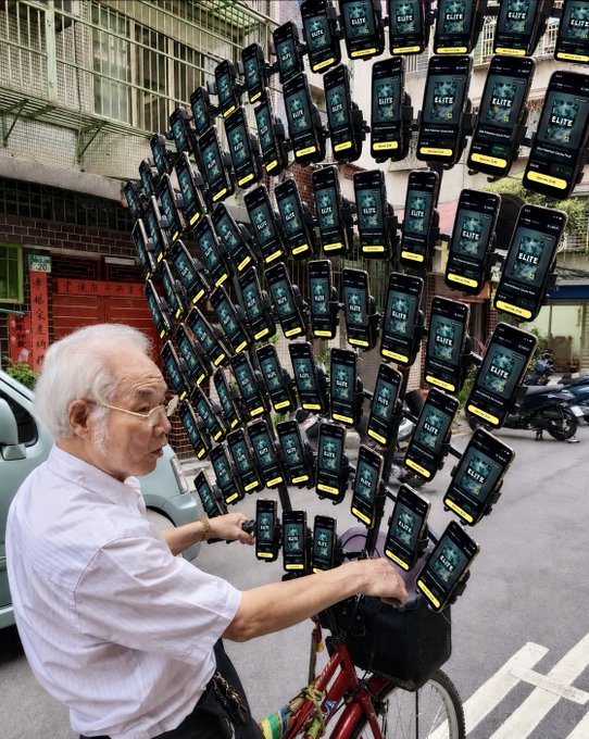 An elderly man rides a red bicycle on a city street while holding an elaborate array of dozens of smartphones displaying 'ELITE' on their screens, all mounted together.