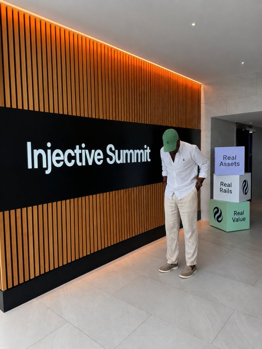 A man in a white shirt and green cap poses in front of an illuminated "Injective Summit" signage at the event in Washington DC, with nearby displays reading "Real Assets," "Real Rails," and "Real Value," reinforcing the summit's focus on advancing on-chain finance.