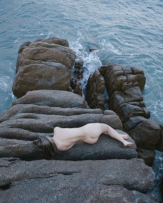 The image features a person lying on their stomach, head resting on a rock, with their body partially submerged in water near the ocean's edge. The photo appears to be taken from an elevated perspective, providing a unique view of the individual against the backdrop of the vast blue sea and the rocky shoreline.