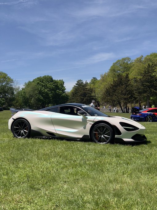 A white McLaren supercar parked on grass at an outdoor car show with trees and a blue sky in the background.