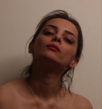 A selfie of a young woman with dark hair, pink lipstick, and a nose piercing against a plain white wall, capturing an intimate close-up portrait.