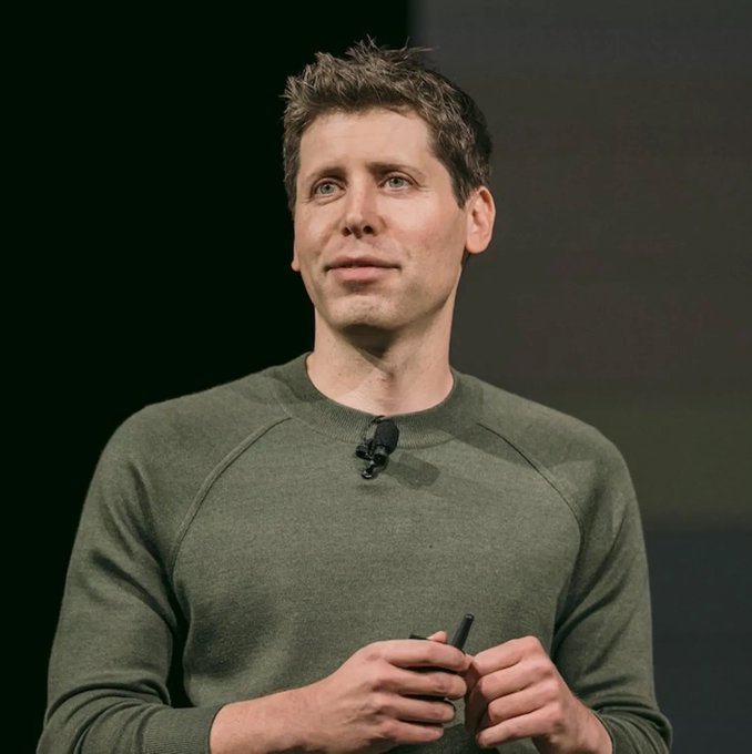 A man with short brown hair wearing an olive green sweater stands on stage with a small lapel microphone, holding a small device in his hands.