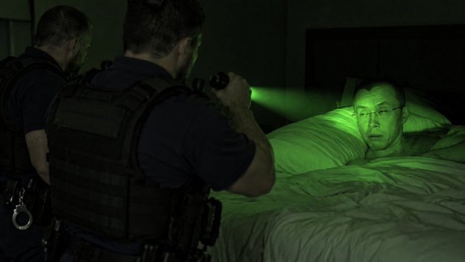 Two armed men in tactical gear are seen from behind, shining a green flashlight at a surprised man wearing glasses who is lying in bed.