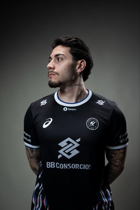 A young man with dark hair and a beard poses in a black athletic jersey featuring "BB Consórcios" as the main sponsor, along with "Serpro" and "Asics" branding, and a circular team crest. The image captures a moment of pride and excitement as he showcases the high-level shirt of his newly joined club, Fluxo.