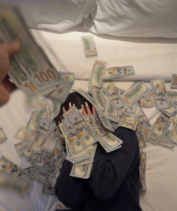 A person lies face-down on a white bed, completely covered and surrounded by scattered $100 US bills, with a hand in the foreground throwing more cash.