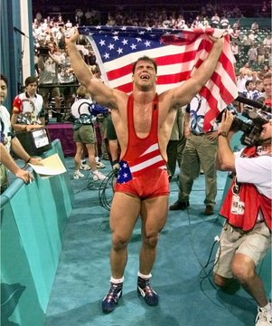 Wrestler Kurt Angle triumphantly holds up an American flag in a crowded arena, wearing a red singlet and smiling in celebration.