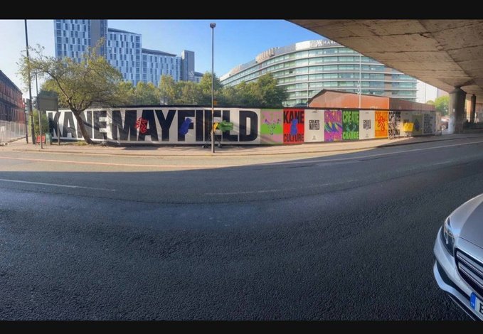 A street view in front of a building displaying the text 'KADEMAYFIELD', featuring colorful murals including one labeled 'KANE CULTURE', with modern buildings and a bridge in the background.