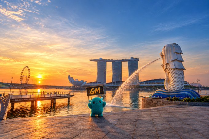 The Pokémon Munchlax holds a sign reading 'GM' in front of Singapore's Marina Bay waterfront at sunset, with the Merlion, Singapore Flyer, and Marina Bay Sands visible in the background.
