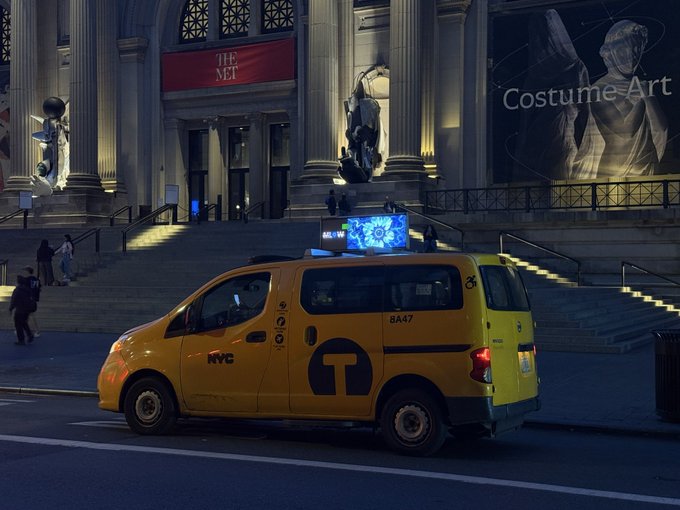 A yellow NYC taxi parked on a street in front of The Met museum at night, which has a "Costume Art" exhibition banner displayed.