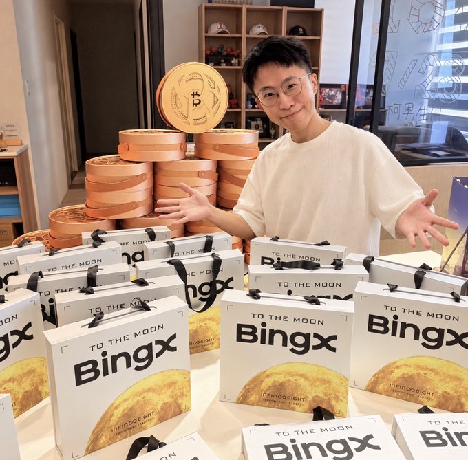 A young man with glasses and a white t-shirt gestures toward stacks of Bingx 'TO THE MOON' branded gift bags and large gold Bitcoin-themed coin props.