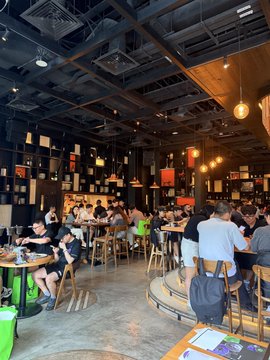 A bustling indoor food court with industrial decor, where many people are seated at high tables and chairs under hanging pendant lights.