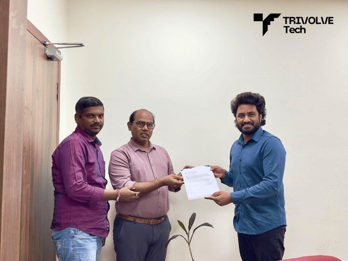 Three men pose indoors, with two exchanging a document under a 'TRIVOLVE Tech' logo.