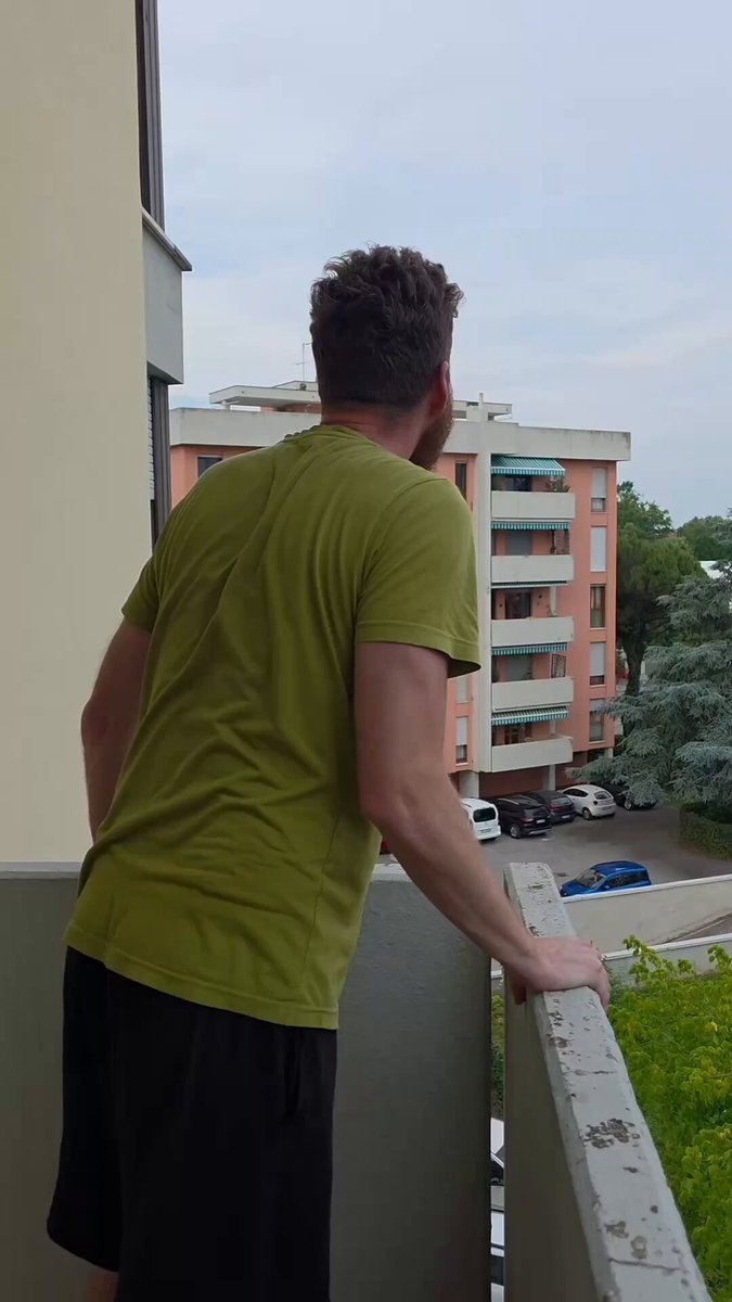 The image shows a man standing on a balcony, looking out at an apartment complex across the street. The balcony has a railing next to him, providing support as he gazes into the distance. There are several cars parked below the apartment building, indicating that this is likely a busy urban area with high pedestrian and vehicular activity.