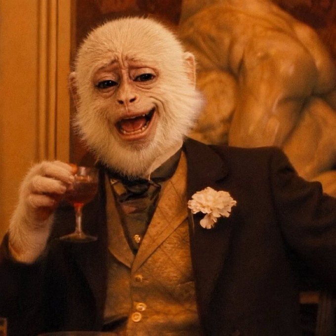 A white monkey wearing a formal suit with a boutonnière holds a small glass of red liquid in an ornately decorated room.