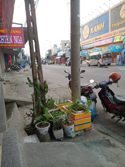 A street scene in Vietnam with shop signs including "Điện Máy XANH" and "thegioid..." visible, with motorbikes parked on the sidewalk next to potted plants.