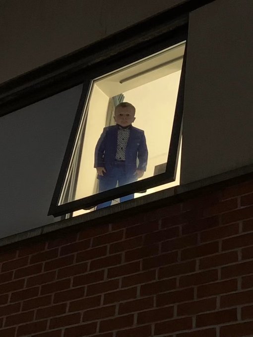 A man in a blue suit and patterned shirt looks out from an open window of a brick building at night, illuminated by interior light.
