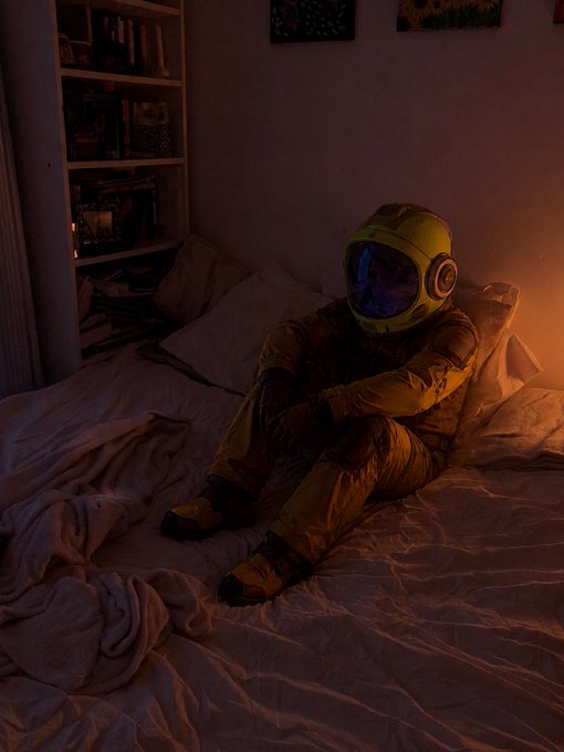 A person in a yellow astronaut suit sits on a bed in a dimly lit room with warm orange lighting.