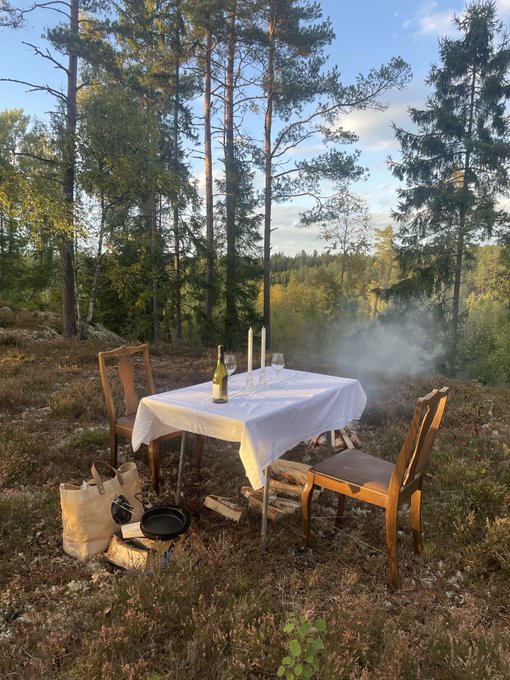 A small table set with a white cloth, wine glasses, and candles sits among trees in a forest clearing next to a smoking fire pit.