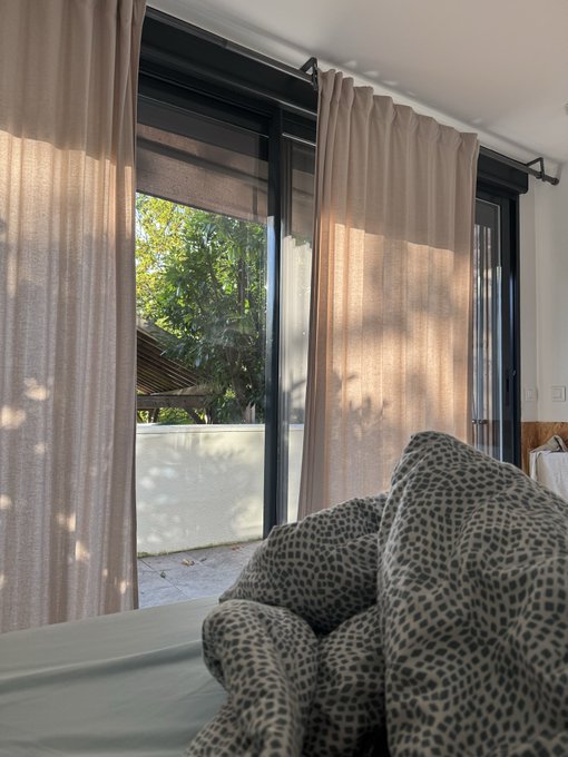 A bright interior view looking out through a window with sheer beige curtains onto a sunlit garden, with a leopard-print blanket or comforter in the foreground on a bed.