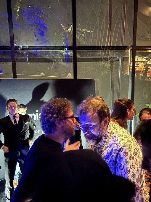 Two men touching foreheads and embracing closely at a dimly lit evening event, with one wearing glasses and a black shirt and the other in a patterned shirt and glasses.