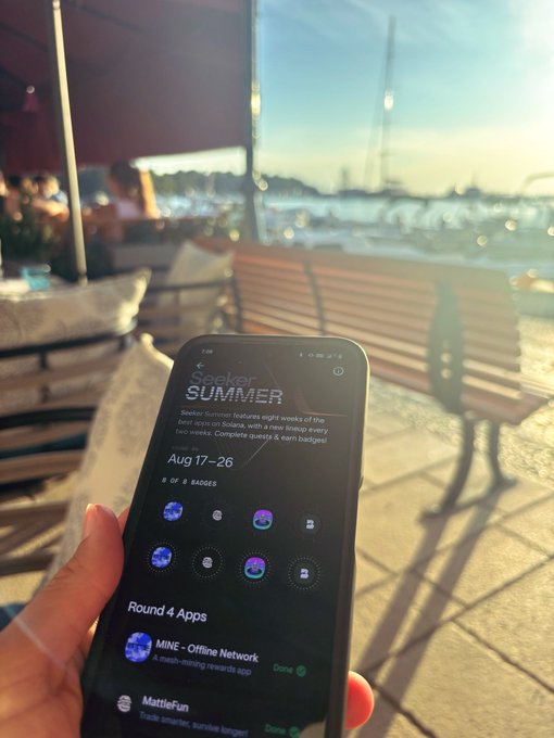 A hand holds a smartphone displaying the "Seeker Summer" app showing "Aug 17-26" with "8 of 8 badges" collected, including "MINE - Offline Network" and "MattieFun" in Round 4, captured in a sunny waterfront setting with a wooden bench and harbor in the blurred background.