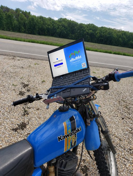 A first-person view from a blue Kawasaki motorcycle parked on a gravel shoulder, with a laptop strapped to the handlebars displaying a login screen for a platform with charts.