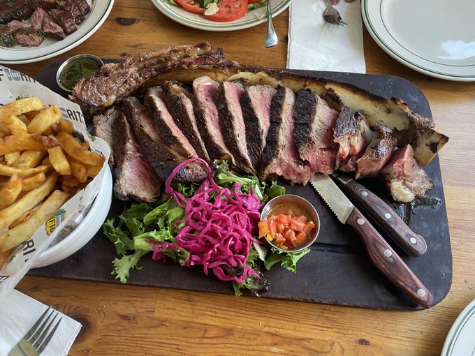 A large wooden board displays a sliced T-bone steak served medium-rare alongside french fries in a paper-lined basket, a vibrant pink pickled onion salad, fresh greens, salsa, and two steak knives.