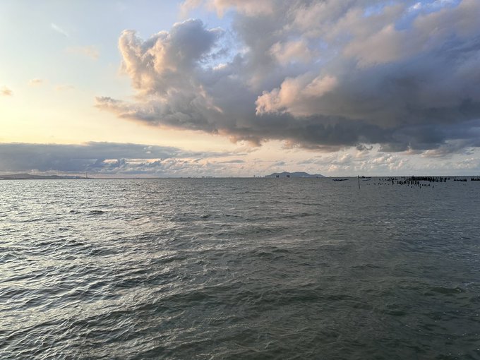 A vast expanse of choppy ocean water stretches beneath a dramatic sky filled with billowing clouds during what appears to be sunset or sunrise. On the right horizon, a small group of people can be seen standing in the shallow water, with a distant coastline visible in the background. The serene seascape perfectly complements the wishes for a peaceful and relaxing weekend.