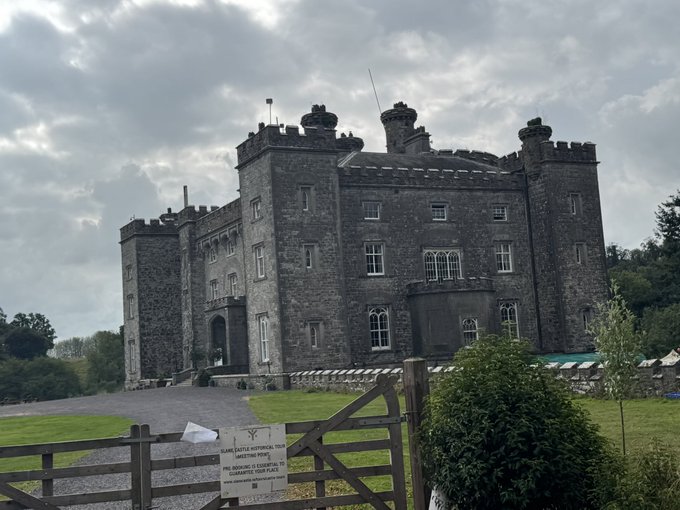 Slane Castle, a historic stone castle in Ireland, stands behind a wooden gate with a sign reading 'Slane Castle Historical Tour' and warnings about no camping, drinking, or firearms on the grounds.
