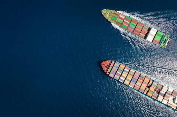 Aerial photograph of two large container ships sailing parallel through deep blue ocean water, carrying stacks of colorful shipping containers.