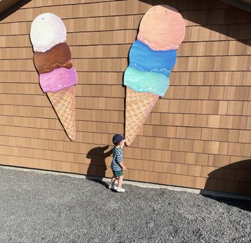 The image features a young boy standing next to two large ice cream cones, one pink and one blue, both adorned with various toppings such as sprinkles and chocolate chips. The boy appears to be looking up at these giant ice cream cones, possibly admiring their size or contemplating which one he would choose if given the option. The scene is set against a backdrop of a brown wall, adding an interesting contrast to the vibrant colors of the ice cream cones.