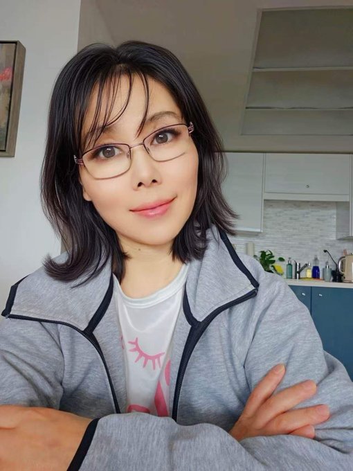 A woman with glasses and dark hair, wearing a gray zip-up jacket over a white shirt, stands with arms crossed in what appears to be a kitchen setting. The image accompanies a Pi2Day message, likely serving as the author's personal photo to connect with fellow Pioneers as she shares insights about the Pi Network vision and ecosystem.