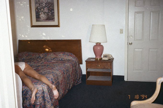 A vintage photograph timestamped '7 18 '94' showing a bedroom scene with only a person's legs visible on a paisley-covered bed, alongside a nightstand with a pink lamp.