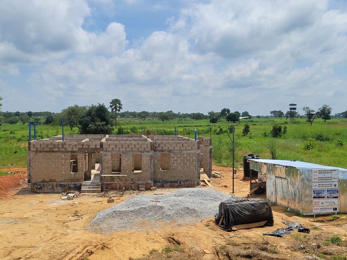 An unfinished brick building under construction sits in a rural landscape, surrounded by a pile of gravel, construction materials, and a small tin-roofed structure, with green fields and trees in the background.