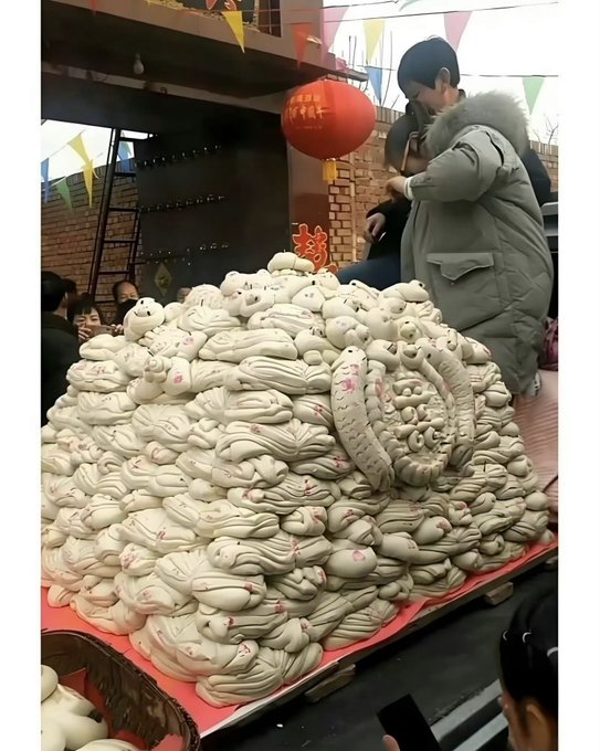 A large pile of intricately shaped white steamed buns (mantou) decorated with red details, arranged on a red table at what appears to be an outdoor market with red lanterns.