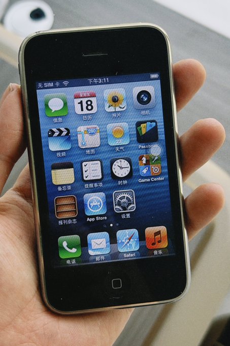 A close-up photo of a hand holding an early-generation iPhone, displaying its home screen with classic iOS app icons including Calendar (showing 18), Photos, Camera, Weather, Maps, Safari, and Music, with the status bar in Chinese reading 下午3:11.