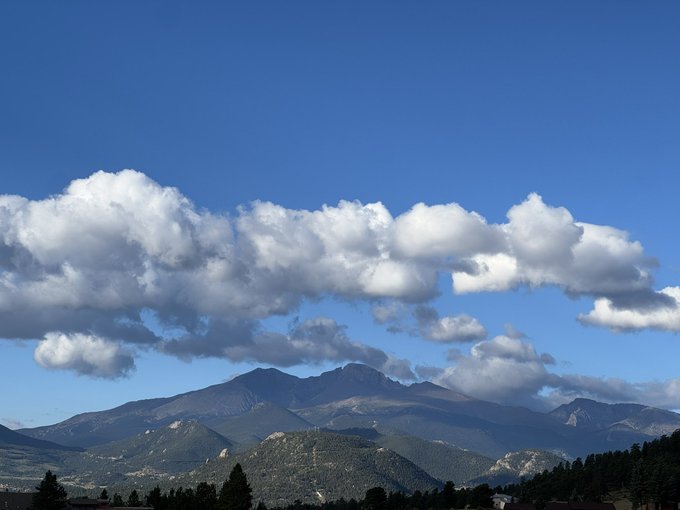 A scenic view of a mountain range with forested hills in the foreground, beneath a bright blue sky filled with fluffy white clouds.