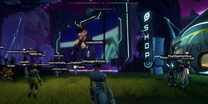 A screenshot from a video game showing multiple player characters gathered in front of a large screen displaying a video of a person wearing a baseball cap, with player names like 'ryancsmith', 'T800-myst', and 'Knuc_ThatSanitariGuy' floating above characters near a structure labeled 'Pulse District'.