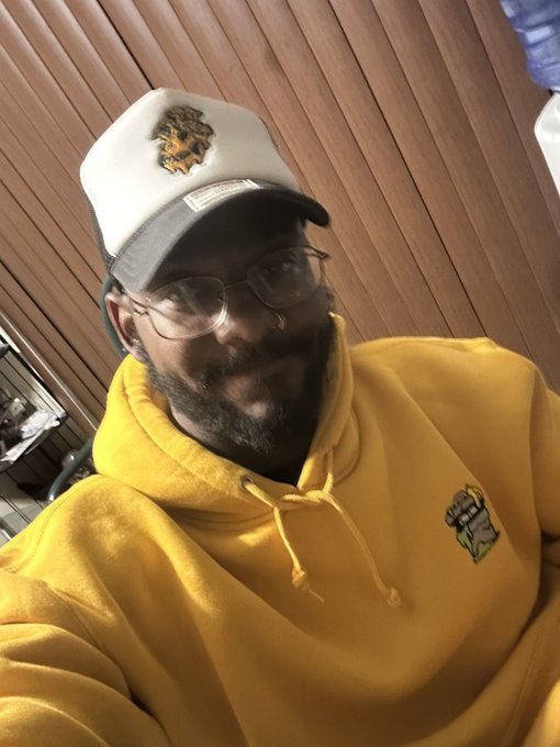 A bearded man wearing glasses, a yellow hoodie featuring an ApeCoin logo, and a trucker hat takes a selfie in front of a wood-paneled wall. The image ties to the #Day561 ApeCoin selfie challenge, showing his support for the $APE token and Yuga Labs community.