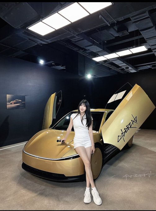 A smiling woman in white poses alongside a gold-colored Tesla Cybercab with its signature butterfly doors open, featuring graffiti-style writing on the interior door panel. The futuristic two-door vehicle is displayed in a modern showroom, fitting the post's reference to the gold Cybercab suiting the setting.