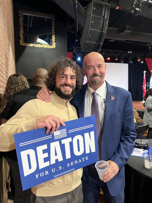 A man with curly hair smiles while holding a "DEATON FOR U.S. SENATE" sign, posing alongside John Deaton, who is dressed in a blue suit and holding a cup at what appears to be a campaign or political event. The image promotes Deaton's upcoming appearance on "Good Evening Crypto" to discuss his Senate campaign, with references to Bitcoin ($BTC) and XRP ($XRP).