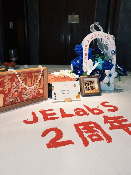 A festive 2nd-anniversary table display for 'JELabs' featuring red text written in granular material, a decorative cake with a card reading 'Wish You Just For You,' blue and white flowers, and a '万事顺利' (Wanshi Shunli) decorative plaque.