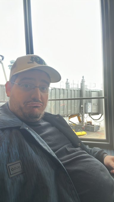 A selfie of a bearded man wearing glasses, a baseball cap, and a dark jacket over a gray shirt, seated indoors with industrial tanks or silos visible through the window behind him.