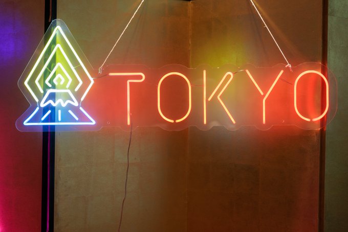 A neon sign displaying 'TOKYO' in red/orange letters next to a multi-colored mountain/volcano logo with green and blue accents, mounted on a wall.