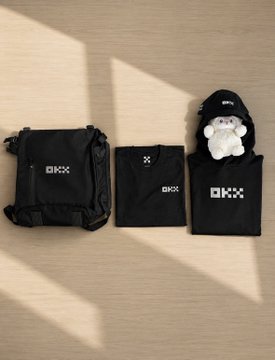 A flat lay photograph of three black OKX-branded merchandise items (a drawstring bag, a small pouch, and a folded item with a plush mascot) arranged on a light wooden floor with warm sunlight casting diagonal shadows.