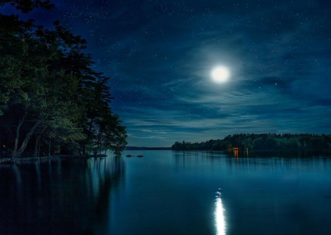 A full moon illuminates a calm lake at night, reflecting on the water with dark trees lining the shore.